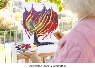 Midsection of caucasian senior woman painting on canvas on sunny terrace. Lifestyle, retirement, senior lifestyle, nature, creativity and domestic life, unaltered. - Powered by Shutterstock