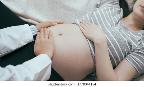 Midsection of female doctor hands careful touching naked belly of pregnant woman in couch at home service living room. asian chinese maternity wife lying on sofa with bare belly having check in house