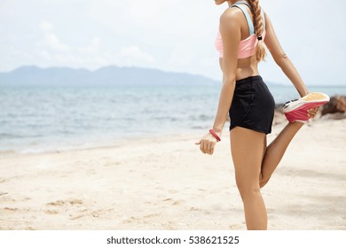 Midsection Of Fit Blonde Sportswoman With Ponytail Warming Up Her Muscles, Stretching Her Legs, Doing Standing Quadricep Front Thigh Stretch Before Running Workout In The Morning, Facing Ocean