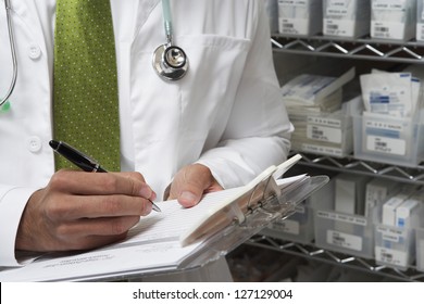 Midsection Of Male Doctor Writing A Prescription While Standing By Medical Supplies On Rack