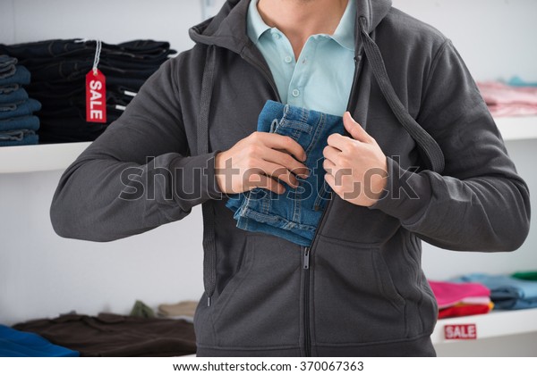 Midsection of man\
hiding jeans in jacket at\
store
