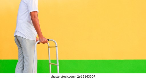 Midsection of middle-aged patient man using walker for support himself in physical walking training in front of colorful cement wall at home, health care concept