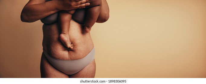 Midsection of mother carrying child while standing against brown background. Postpartum belly with stretch marks.