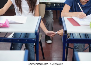 Midsection Of Schoolboy Passing Cheat Sheet To Girl During Exam At Desk In Classroom