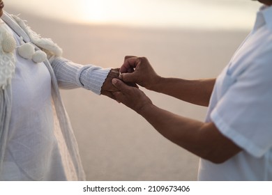 Midsection of senior biracial groom putting wedding ring on bride's finger at beach during sunset. lifestyle, culture and love. - Powered by Shutterstock