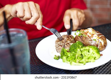 Midsection Of Senior Man Eating Strip Steak Served With Loaded Baked Potato And Broccoli At Restaurant Table