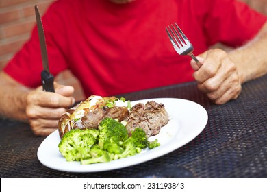 Midsection Of Senior Man Having Strip Steak Served With Loaded Baked Potato And Broccoli At Restaurant Table