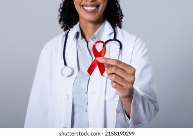 Midsection of smiling african american mid adult female doctor holding red aids awareness ribbon. white background, hiv, aids, doctor, medical, disease, awareness, support and healthcare concept.