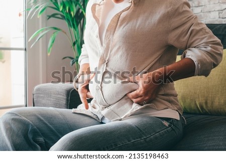Midsection of woman hands holding her belly fat sitting on sofa at home. Caucasian lady grabbing excessive fat on her abdomen. Young female pinching on her fatty obese waist