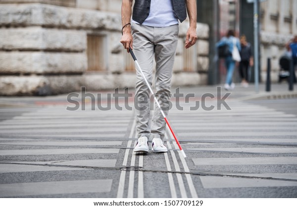Midsection of young blind man with white cane\
walking across the street in\
city.