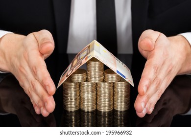 Midsection of young businessman protecting house made of currency at desk
