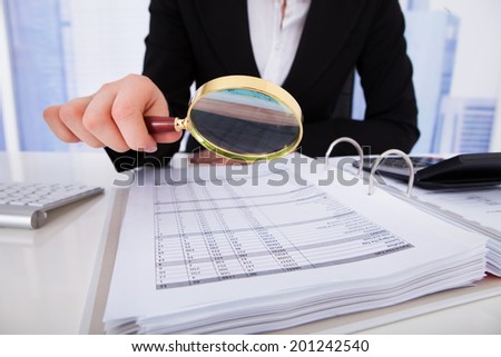 Midsection of young businesswoman scrutinizing bills with magnifying glass at office desk