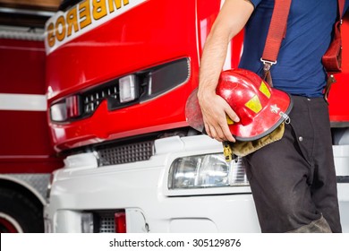 Midsection Of Young Fireman Holding Red Helmet While Leaning On Firetruck At Station