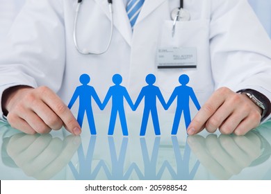 Midsection Of Young Male Doctor Holding Blue Paper People Chain At Desk In Clinic