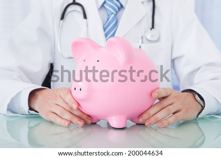 Midsection of young male doctor holding piggybank at desk in clinic