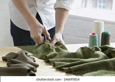 Midsection of young male tailor cutting piece of cloth at table in fashion studio