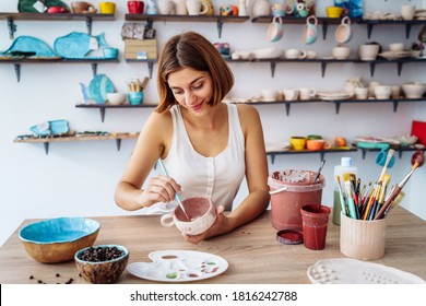 Mid-shot of potter decorating clay mug after firing in oven. Woman in white tanktop enjpying creative procces of pottery coloring. Sitting in pottery workshop with white walls and lorful clay products