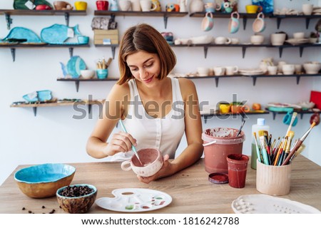 Mid-shot of potter decorating clay mug after firing in oven. Woman in white tanktop enjpying creative procces of pottery coloring. Sitting in pottery workshop with white walls and lorful clay products