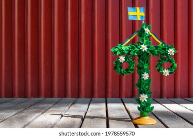 A midsummer pole with flag against red wooden wall. Handmade maypole decorated with flowers and leaves for celebrating the midsummer day. Kort Glad Midsommar in Swedish or Card Happy Midsummer in en