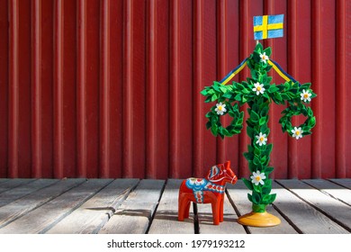 A midsummer pole with flag and Dala horse against red wooden wall. Handmade maypole decorated with flowers for celebrating midsummer day. Kort Glad Midsommar in Swedish or Card Happy Midsummer in eng. - Shutterstock ID 1979193212