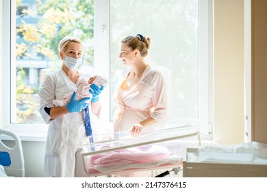 Midwife or doctor with newborn concept. Woman nurse doctor or midwife in medical mask standing holding newborn baby infant in hands and young mother in front of the window.