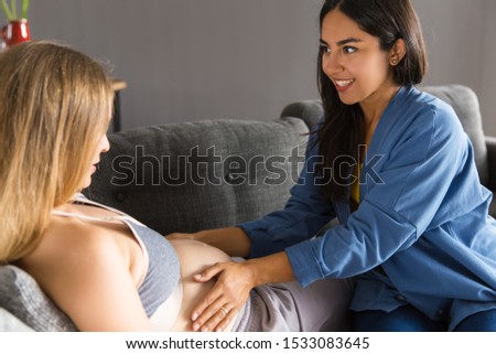 Midwife examining expectant mother at home. Young woman touching belly of pregnant patient lying on couch. Pregnancy and healthcare concept