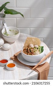 Mie Ayam Bangka Or Noodle With Bean Sprout, Meatballs, Crackers And Chicken Cubes, Served On White Bowl. Cafe Table Corner Situation With Some Indoor Plant. Popular Indonesian Street Food. 