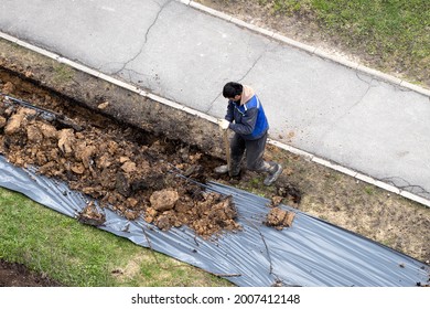 Migrant Labor. A Worker Digging A Ditch Along A Pavement In Summer.