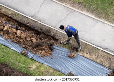 Migrant Labor. A Worker Digging A Ditch Along A Pavement In Summer.