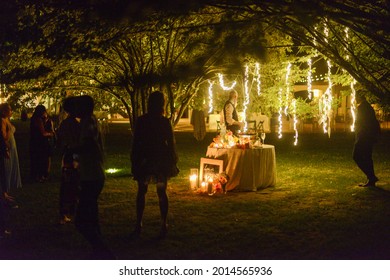 MILA, ITALY - Jul 07, 2021: A Small Round Table At A Wedding Venue In A Park At Night With People Gathered Around It For The Cake Cutting