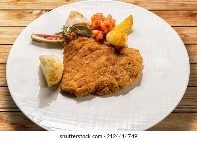 Milanese Cutlet, Veal Cutlet With Bone Breaded And Fried In Butter. In White Dish With Potatoes And Tomatoes On Wooden Table