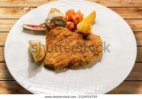 Milanese\
cutlet, veal cutlet with bone breaded and fried in butter. In white\
dish with potatoes and tomatoes on wooden\
table