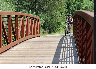 Milford, MI/USA: July 29, 2017 – Woman Looking At Cell Phone Casually Bicycles Over Footbridge On The Huron River Water Trail, A 104 Mile Inland Paddling Trail In The Lower Peninsula Of Michigan. 