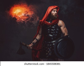 Military bearded roman in red hood and dark armor which holding an axe and shield posing in studio room with spotlight and smoke.