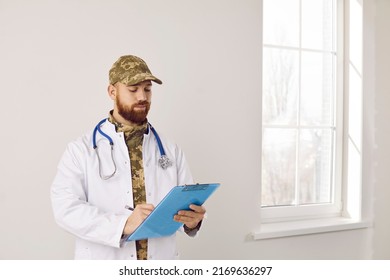 Military Doctor Inside The Hospital. Man With Ginger Beard, Wearing Camouflage Uniform And White Medical Lab Coat Holding Clipboard And Reading Some Papers. Medicine And Healthcare In The Army Concept