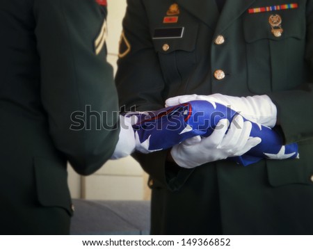 Military honor guard carefully folds the United States flag for presentation to family members at a veteran's funeral. Selective focus on flag and gloved hands. 