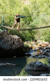 A Military Man Or Airsoft Player In A Camouflage Suit Sneaking The Rope Bridge And Aims From A Sniper Rifle To The Side Or To Target. 