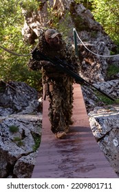 A Military Man Or Airsoft Player In A Camouflage Suit Sneaking The Rope Bridge And Aims From A Sniper Rifle To The Side Or To Target. 