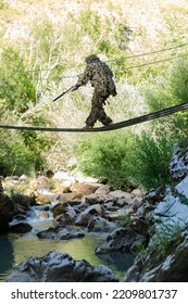 A Military Man Or Airsoft Player In A Camouflage Suit Sneaking The Rope Bridge And Aims From A Sniper Rifle To The Side Or To Target. 