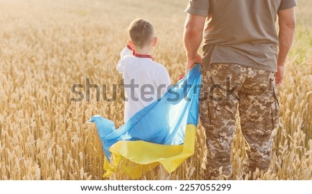 Military man and child with Ukraine flag in wheat field. Ukraine independence day concept. Stop war in Ukraine. Save Ukraine