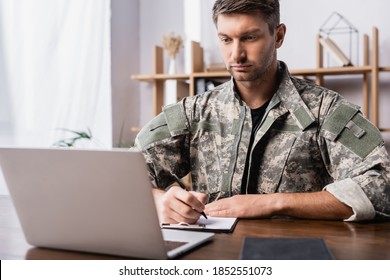 Military Man In Uniform Holding Pen While Writing On Clipboard Near Laptop On Desk