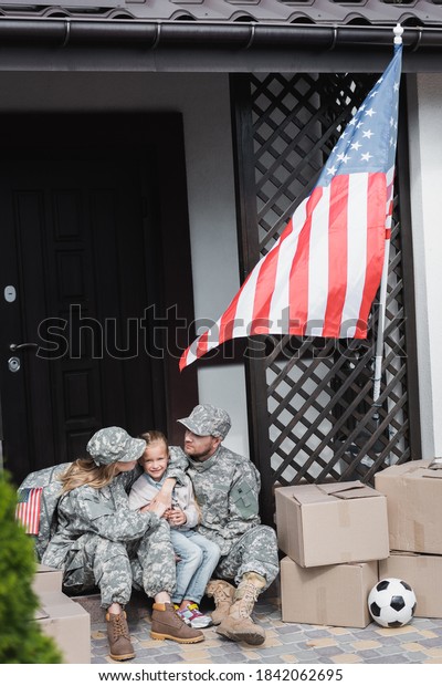 Military parents with daughter hugging, while\
sitting on threshold near cardboard boxes and american flag on\
blurred foreground