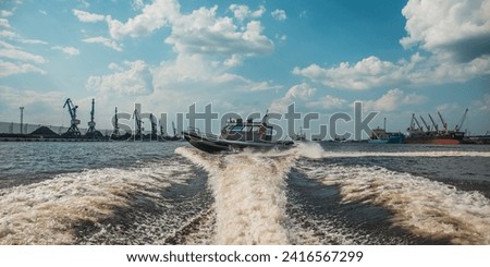 military patrol boat on a mission in a harbor with industrial cranes and cargo ships in the background under a cloudy sky
