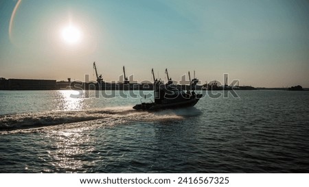military patrol boat on a mission in a harbor with industrial cranes and cargo ships in the background under a cloudy sky