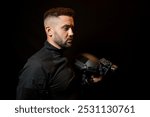 Military, policeman of European appearance with a beard in a black uniform with a tactical helmet with a night vision device in his hand on a black background.