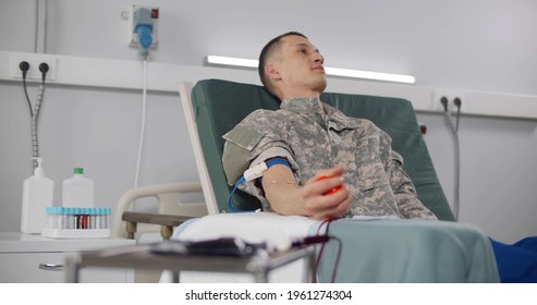 Military Servant Volunteer During Donation With Squeezing Red Rubber Heart In Hand. Portrait Of Soldier In Chair Donating Blood In Medical Center Or Hospital