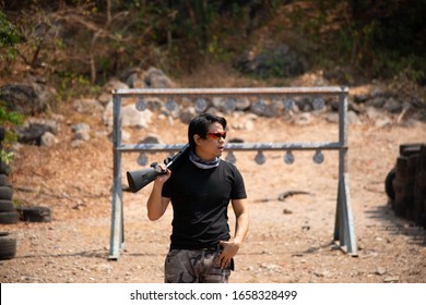 Military Sport Man Short Hair Wear Solid Black Shirt, Camoflage Plant, Camouflage Scarf And Red Dark Shade Holding Black Shotgun On Right Shoulder And Loking On Left Standing In Shooting Range