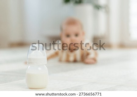 Milk bottle in sharp focus with crawling baby in striped pajamas blurred in background, reaching forward on soft indoor floor.