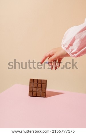 Milk chocolate on a beige-pink background. The girl's hand reaches for the chocolate. Creative chocolate photography. Space for text. Vertical