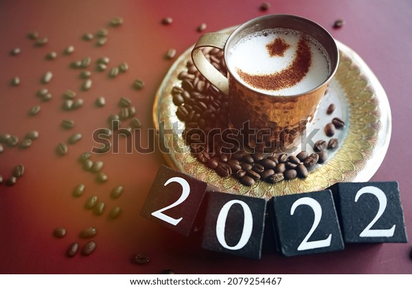 Milk or coffee cup with star and crescent moon\
sign over frothy surface served on red background with wooden cubes\
number 2022 and coffee beans. Ramadan food and drink, iftar meal\
for the holy month.